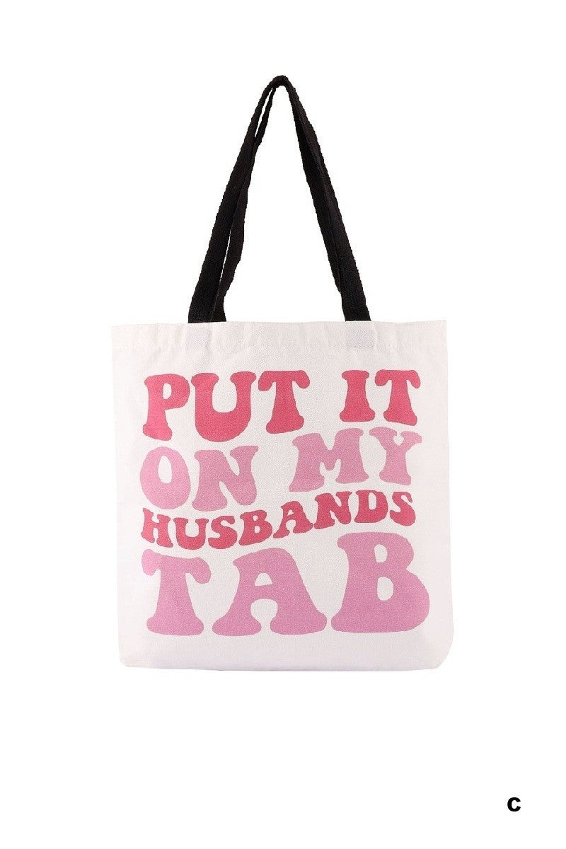Tote bag with pink text 'PUT IT ON MY HUSBAND'S TAB' on a white background