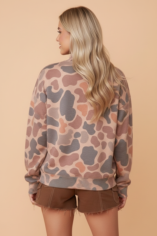 A woman with long blonde hair is shown from the back, wearing a long-sleeve crewneck sweatshirt with a muted camouflage pattern in dusty pink, brown, and gray, paired with brown shorts, against a solid beige background.