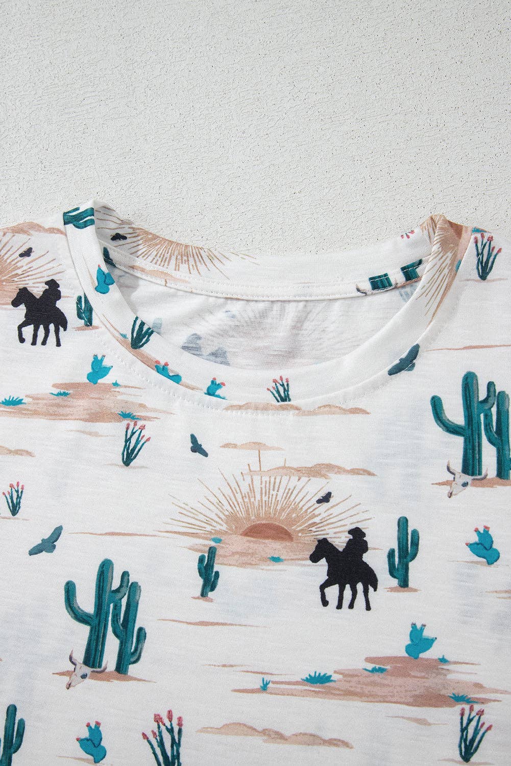 White shirt with desert-themed design featuring cacti, horses, and sunsets on a white background.