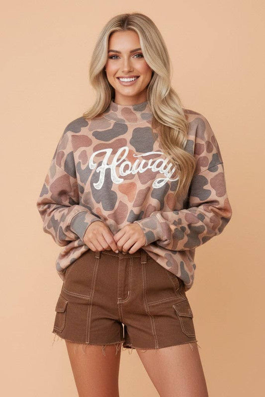 A woman modeling a brown and beige camouflage sweatshirt with 'Howdy' written in white script across the chest, paired with brown cargo shorts against a solid peach background.